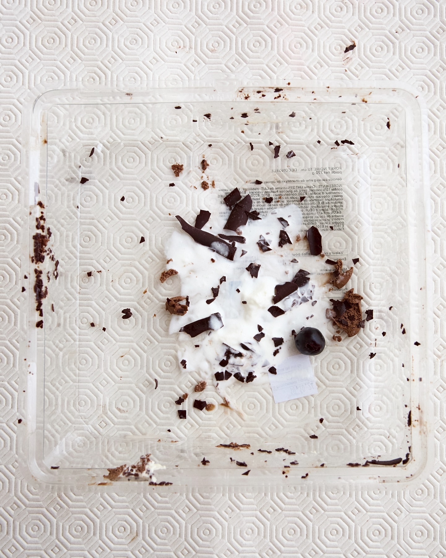 Overhead shot of a clear square plastic container with leftover cake crumbs, white cream, pieces of dark chocolate, and a single berry.