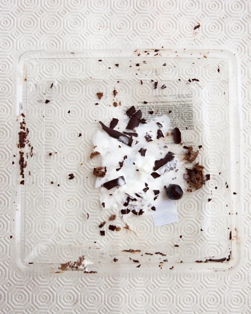 Overhead shot of a clear square plastic container with leftover cake crumbs, white cream, pieces of dark chocolate, and a single berry.