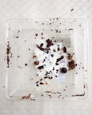 Overhead shot of a clear square plastic container with leftover cake crumbs, white cream, pieces of dark chocolate, and a single berry.
