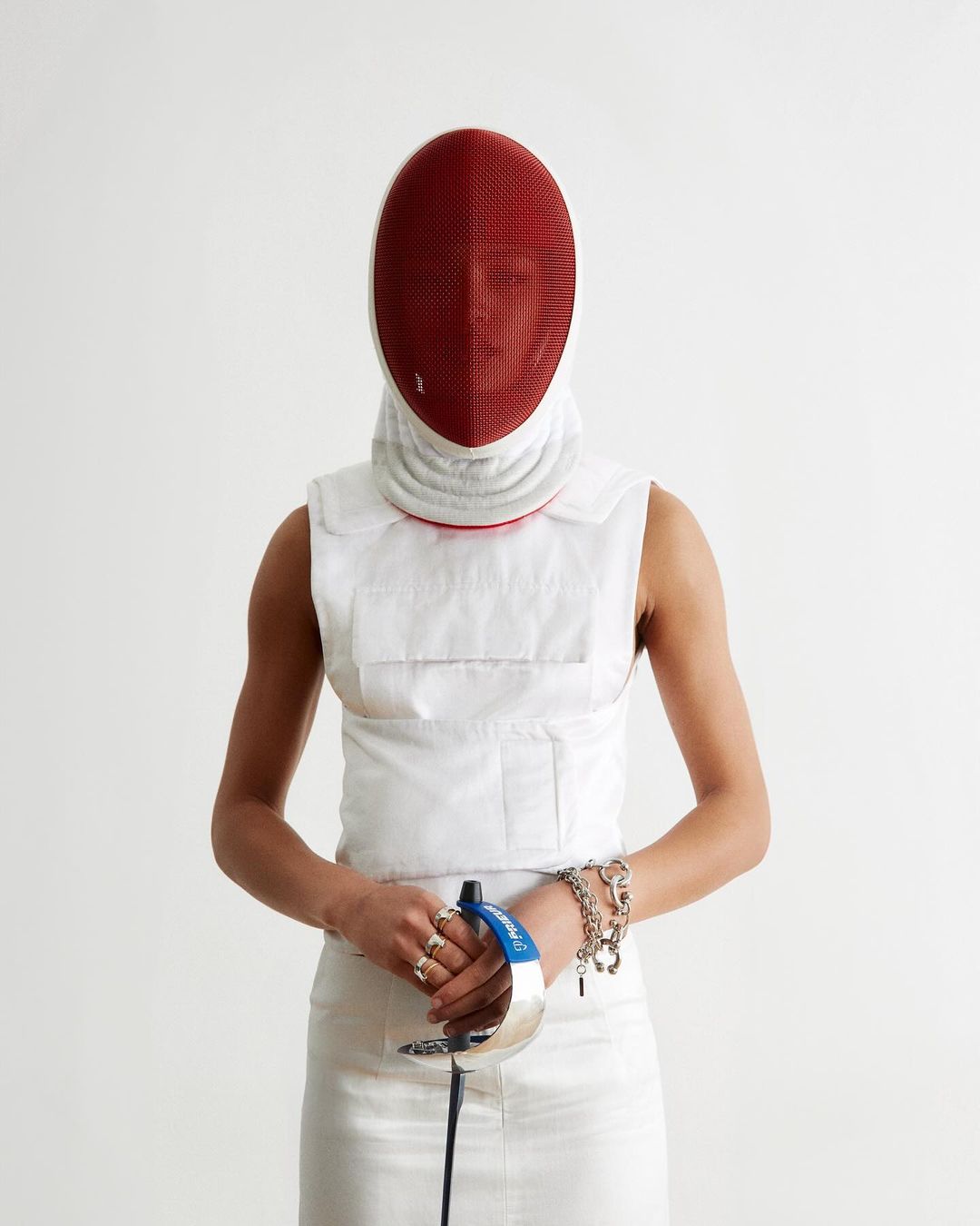 A fencer in white uniform and red mask stands ready with foil in a white studio, showcasing sport and protective gear.