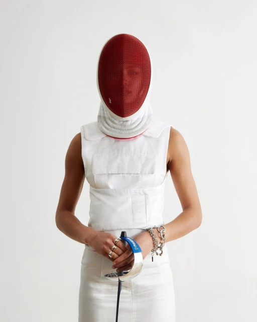 A fencer in white uniform and red mask stands ready with foil in a white studio, showcasing sport and protective gear.