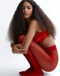 A woman with long curly brown hair crouching and wearing vibrant red underwear and tights against a seamless white background.