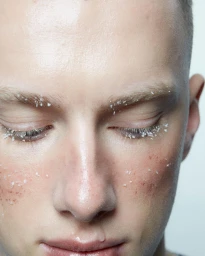 Close-up portrait of a young man with closed eyes, frosted eyelashes, and tiny crystal flakes scattered across his face.