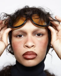 Close-up portrait of a model with dark glossy lips holding tinted sunglasses above messy dark hair against a white background.