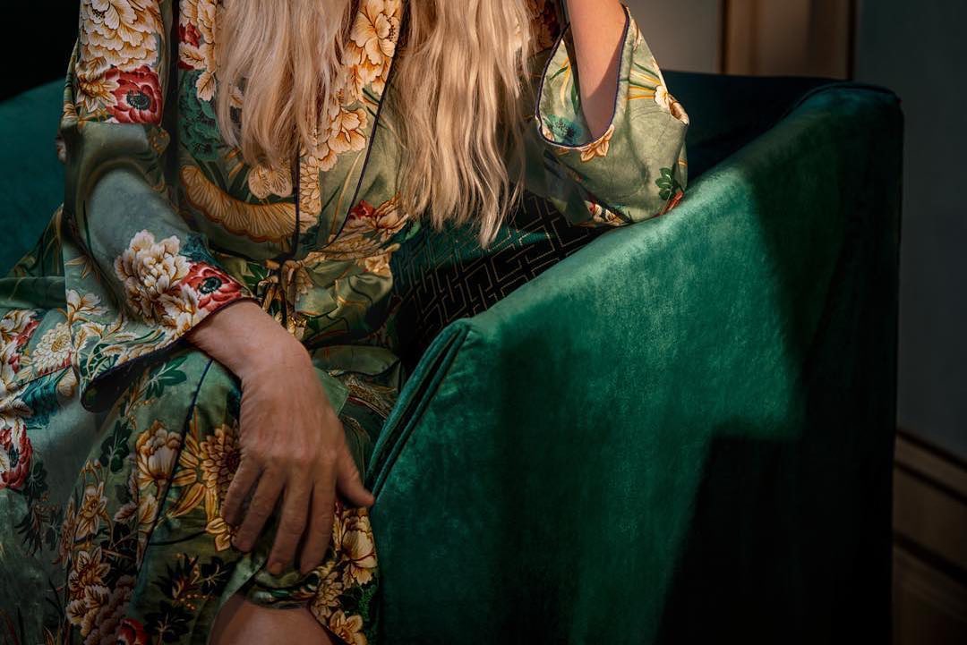 Woman with long blonde hair sits in a green velvet chair, wearing a green floral robe in an indoor setting with subdued lighting.