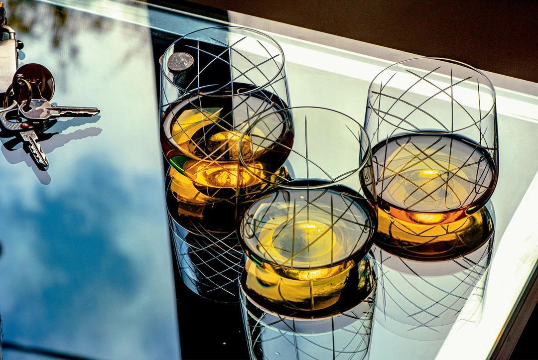 Three patterned glasses, two partially filled with amber liquid, on a reflective surface next to keys. Indoor setting, cool lighting.