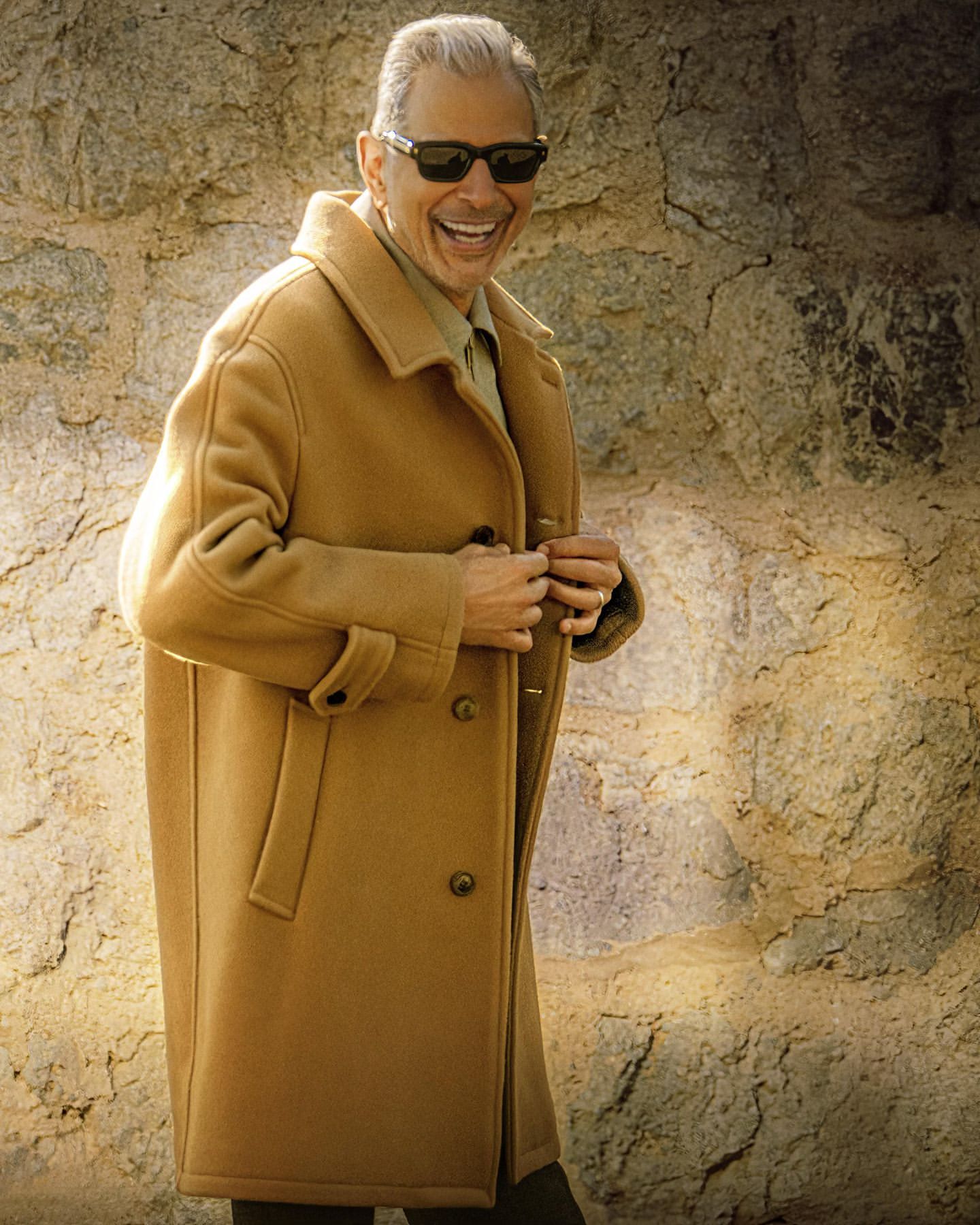 Portrait of a smiling, mature adult male wearing sunglasses and a tan overcoat, standing outdoors against a textured stone wall.