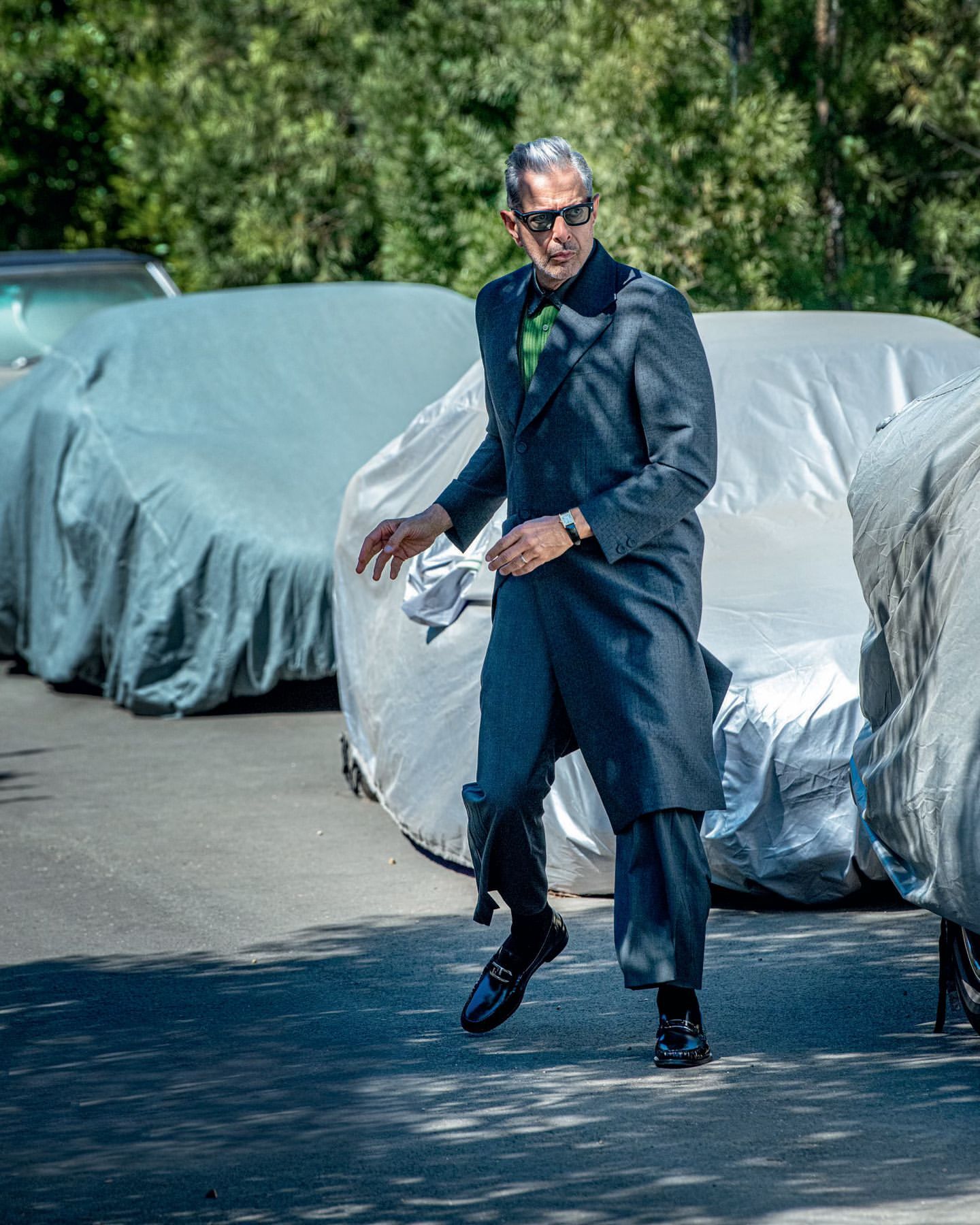 Jeff Goldblum in a grey coat and black loafers walks outside on pavement near parked, covered cars on a sunny day.