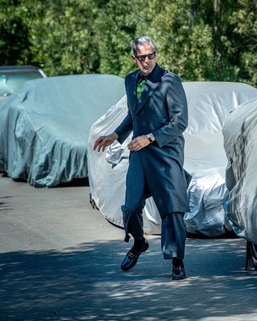 Jeff Goldblum in a grey coat and black loafers walks outside on pavement near parked, covered cars on a sunny day.