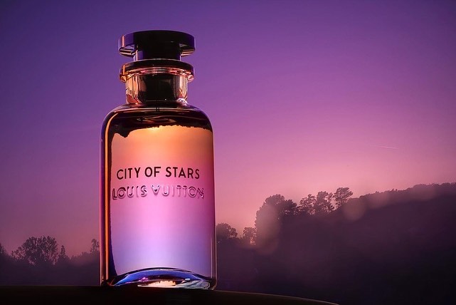 Elegant bottle of Louis Vuitton 'City of Stars' perfume is set against a dreamy sunset background with hints of forest trees.