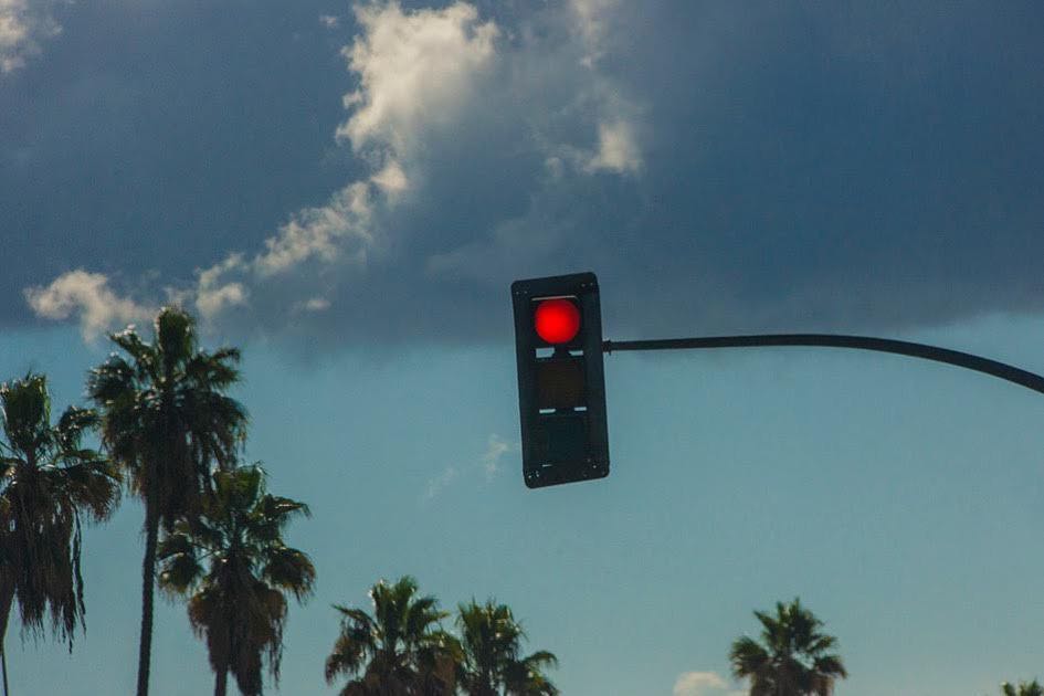 Red traffic light against a cloudy sky with palm trees, signalling 'stop'. Transportation safety visual with clear signal.