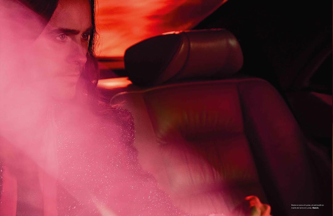 An adult male sits in the red leather seat of a car illuminated with red lighting, captured in an editorial fashion style.