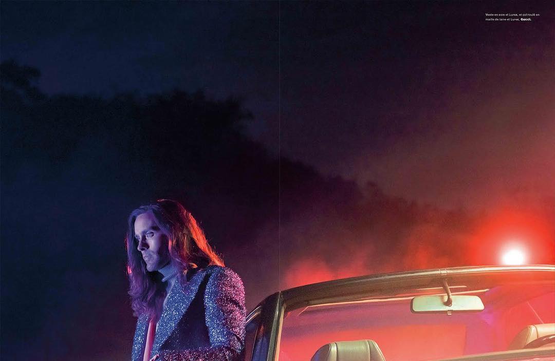 Adult male with long hair wearing a patterned jacket standing next to a convertible car at night with dark clouds.