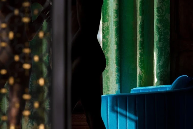 Silhouette of a person framed by a curtain with a floral pattern and an upholstered blue chair against a dark background.