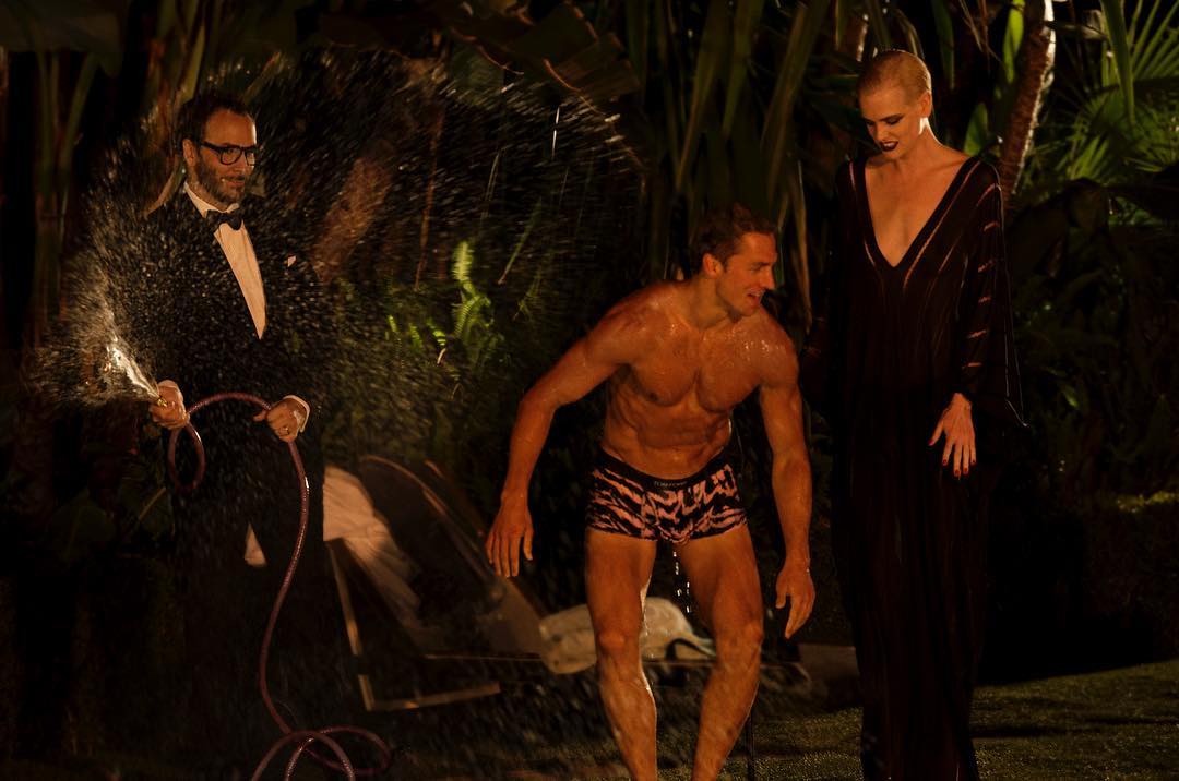 Man in tuxedo sprays water on shirtless man and woman in black dress during an outdoor event at night with plants in the background.