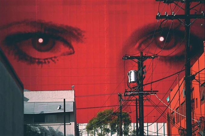 Urban mural featuring large eyes painted on a red building wall, with power lines and adjacent buildings adding depth to the scene.