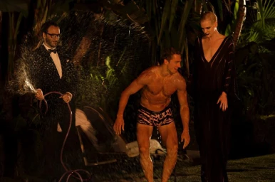 Man spraying water from a hose next to a shirtless man in printed briefs and a woman in a long black dress at night outdoors.