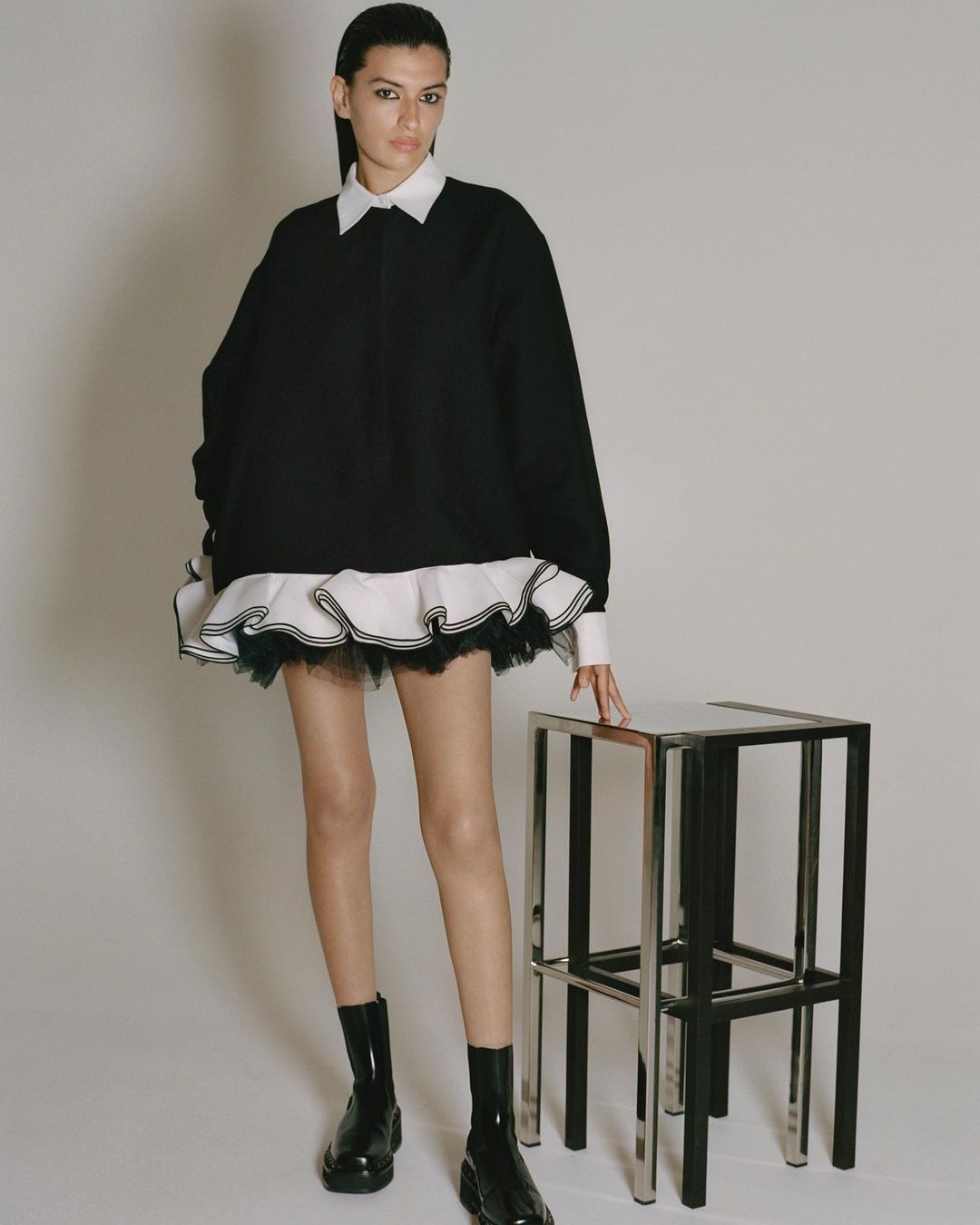 Full-length shot of a slender female model in a black sweater dress with a white collared shirt, standing next to a silver stool.