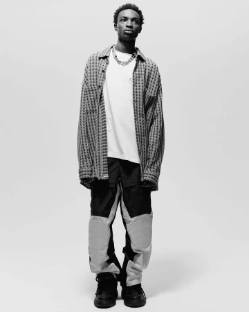 Black man standing in a plaid overshirt, white t-shirt, and colour-blocked trousers against a plain white background.
