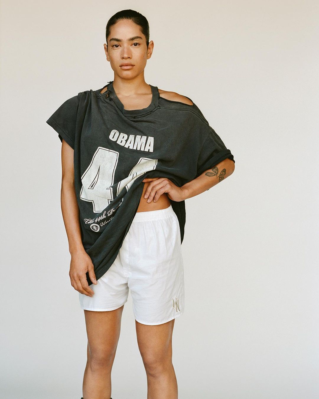 A young adult female poses in a studio, wearing an oversized distressed t-shirt and white boxer shorts against a neutral backdrop.