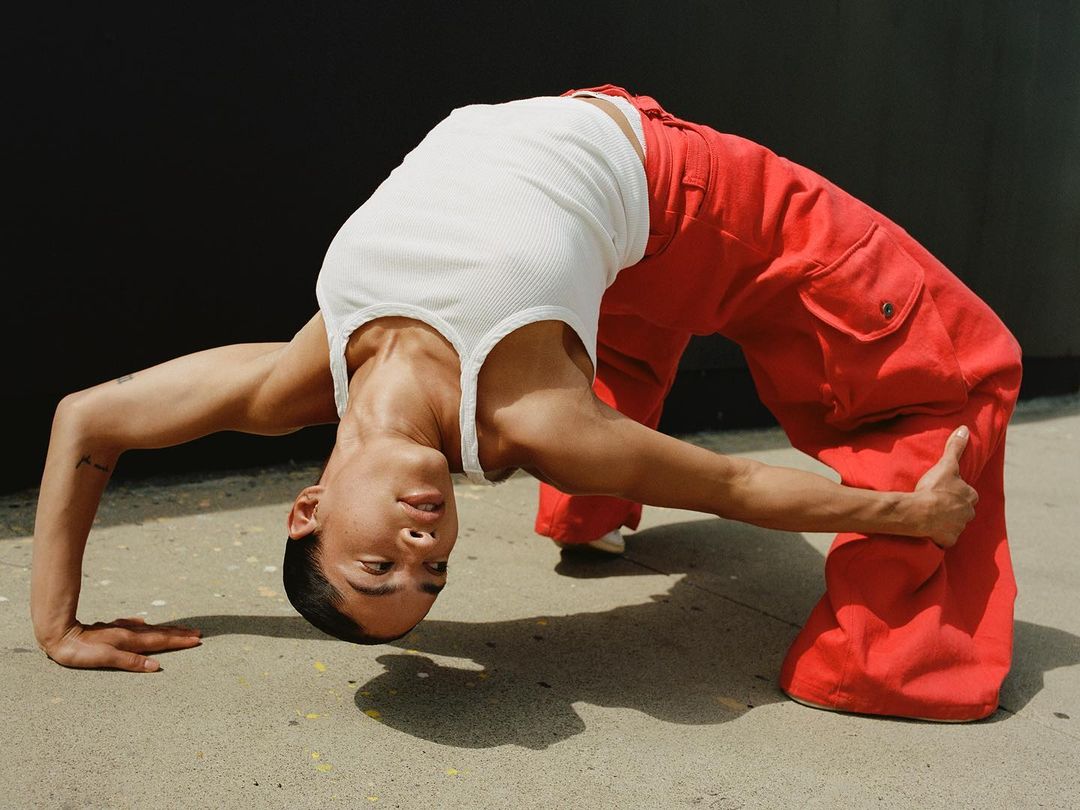 Adult man in white ribbed tank top and red pants, in a backbend pose on a city sidewalk, bending over backwards with hands on ground.