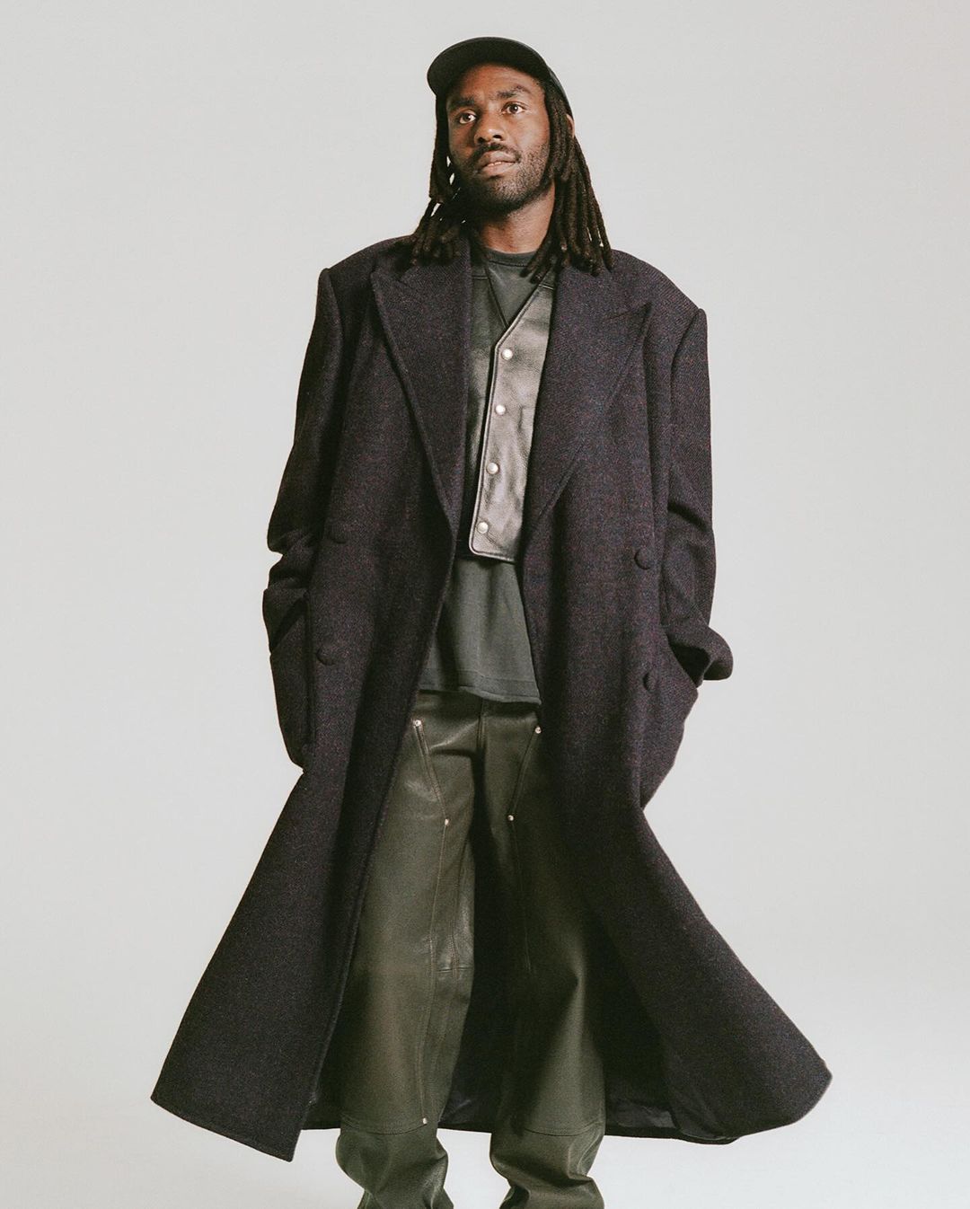Black man with dreadlocks wearing a long dark wool coat, dark cap, and leather vest against a plain grey background