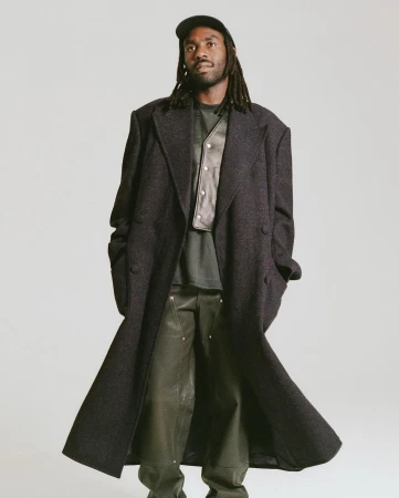 Black man with dreadlocks wearing a long dark wool coat, dark cap, and leather vest against a plain grey background