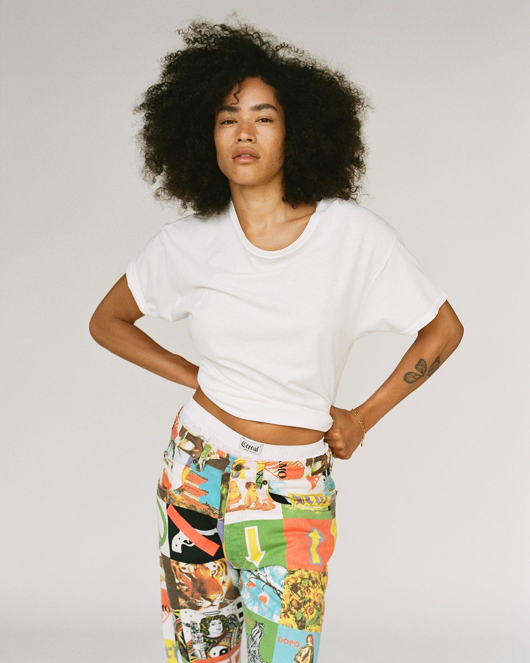 An adult female stands in a photo studio, wearing a white t-shirt and patchwork pants. She is photographed in medium shot.
