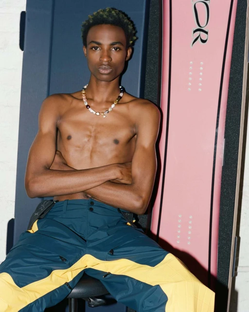 Shirtless young adult poses indoors with a light pink snowboard and wears colorful pants and necklace.
