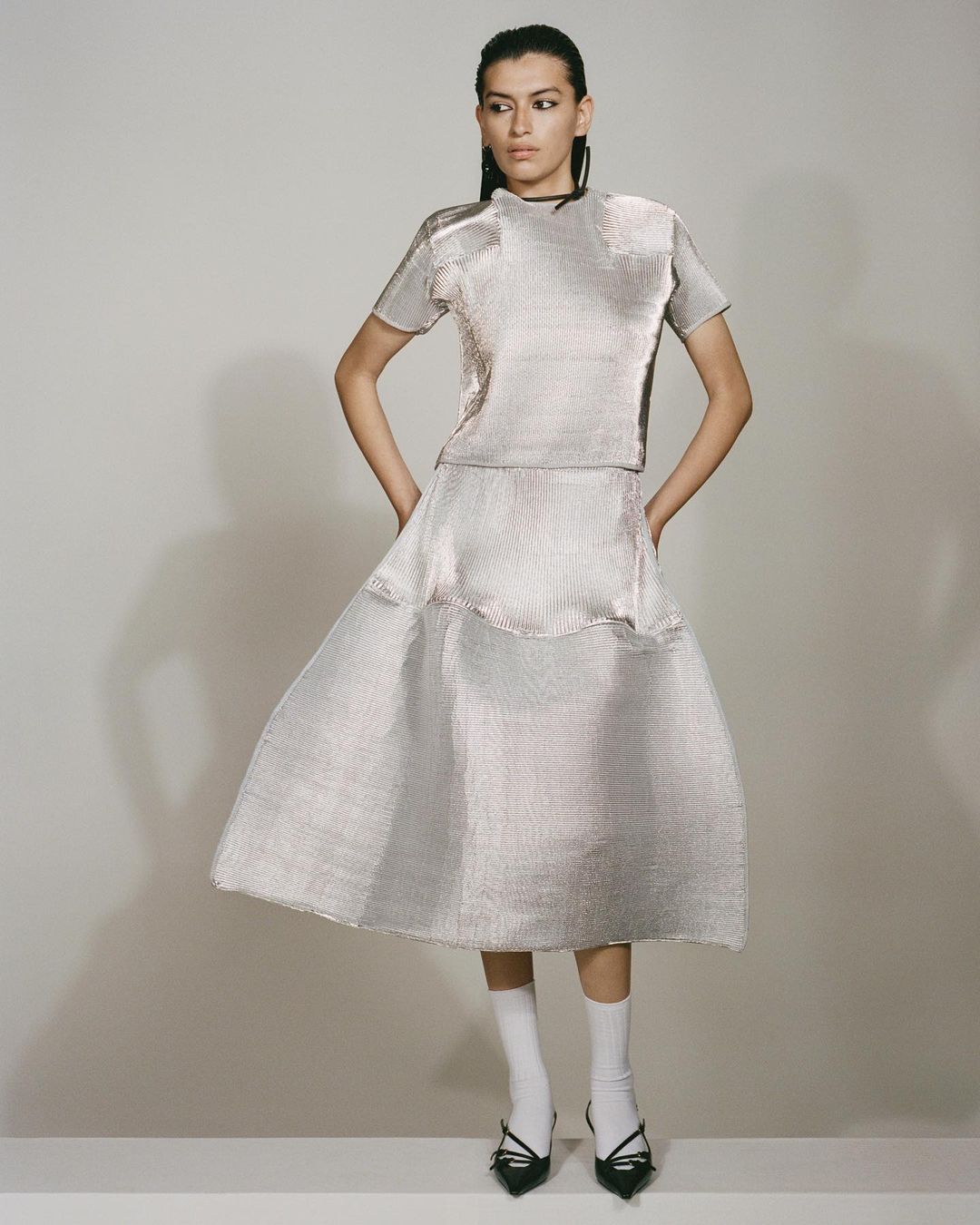 A woman in a metallic silver top and skirt stands against a neutral background, wearing white socks and black shoes for a fashion-forward look.