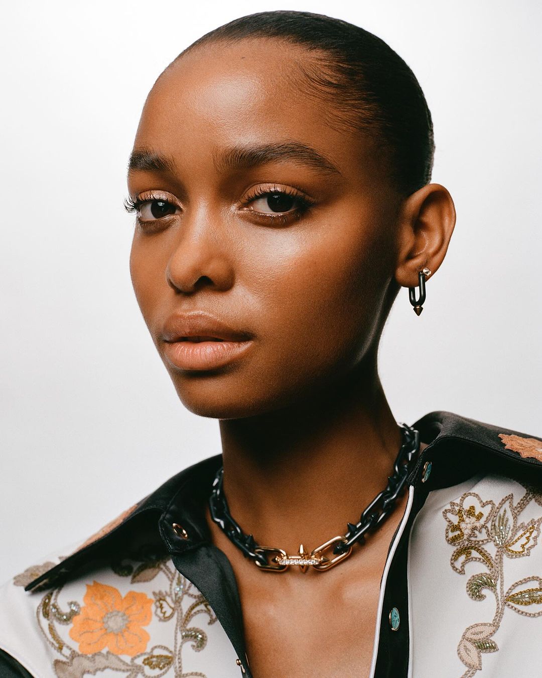 Close-up portrait of a Black woman wearing a patterned floral shirt, chunky chain necklace, and small earrings against a plain white background.