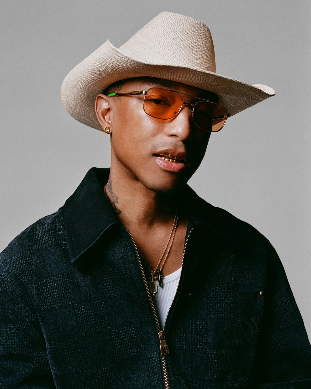 Portrait of Pharrell Williams wearing a cowboy hat and orange aviator sunglasses in front of a neutral studio backdrop.