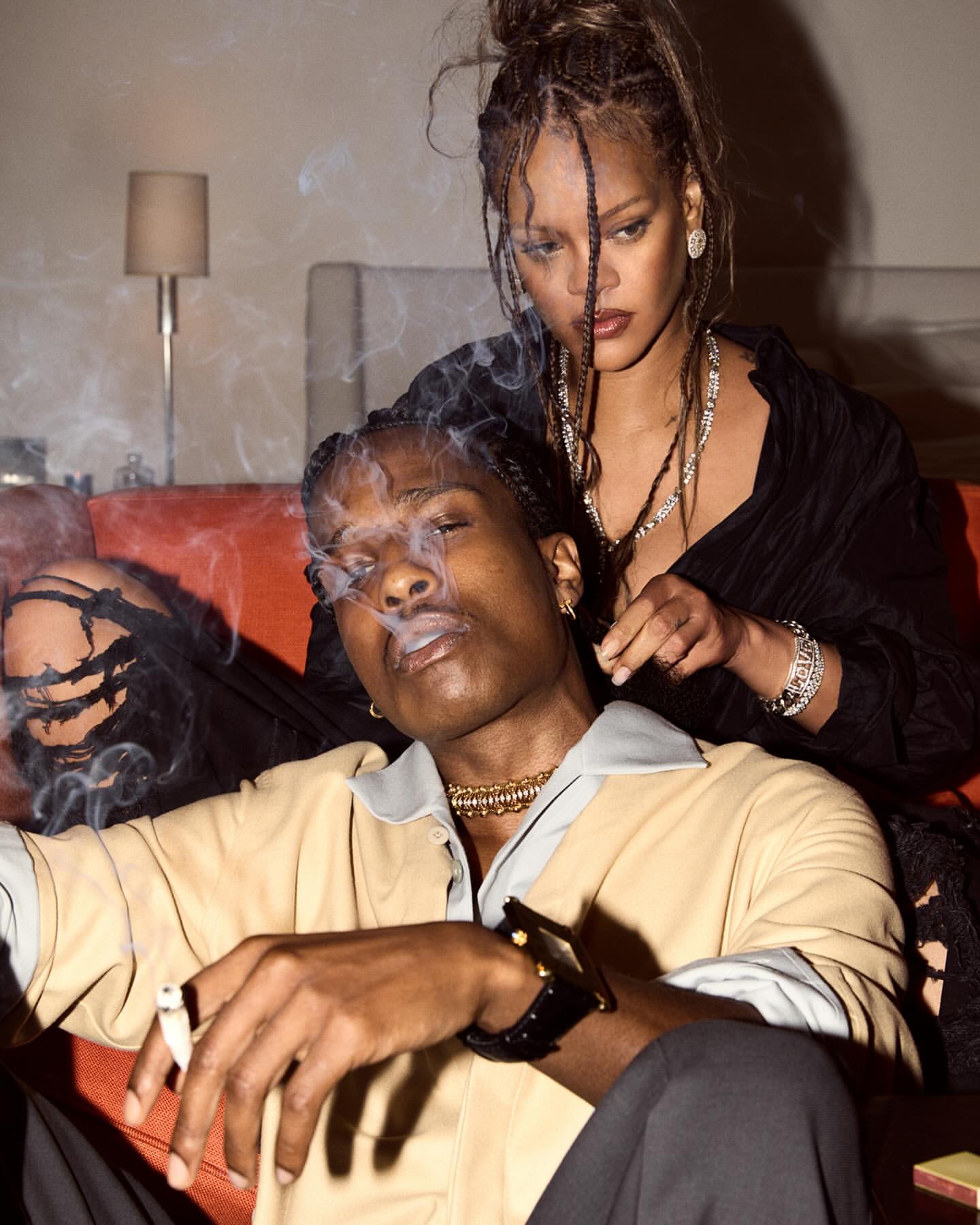 Portrait of A$AP Rocky smoking with Rihanna behind him, both wearing jewelry and stylish clothes, in an indoor setting with soft lighting.