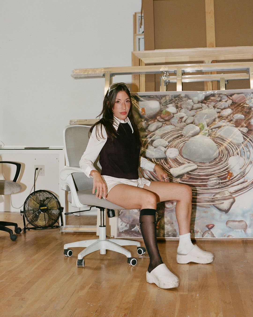 Young woman in black vest and white top sits on rolling chair in bright room with artwork. She is wearing sheer black socks.