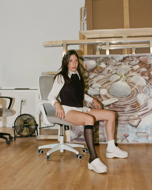 Young woman in black vest and white top sits on rolling chair in bright room with artwork. She is wearing sheer black socks.