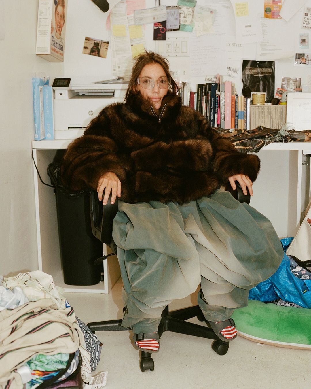 A woman with glasses in a fur coat sits in an office chair surrounded by objects in an indoor setting with natural lighting.