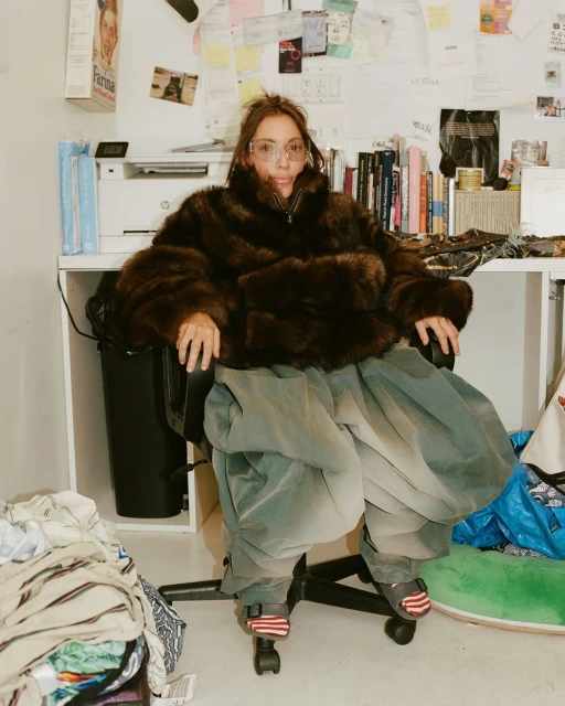A woman with glasses in a fur coat sits in an office chair surrounded by objects in an indoor setting with natural lighting.