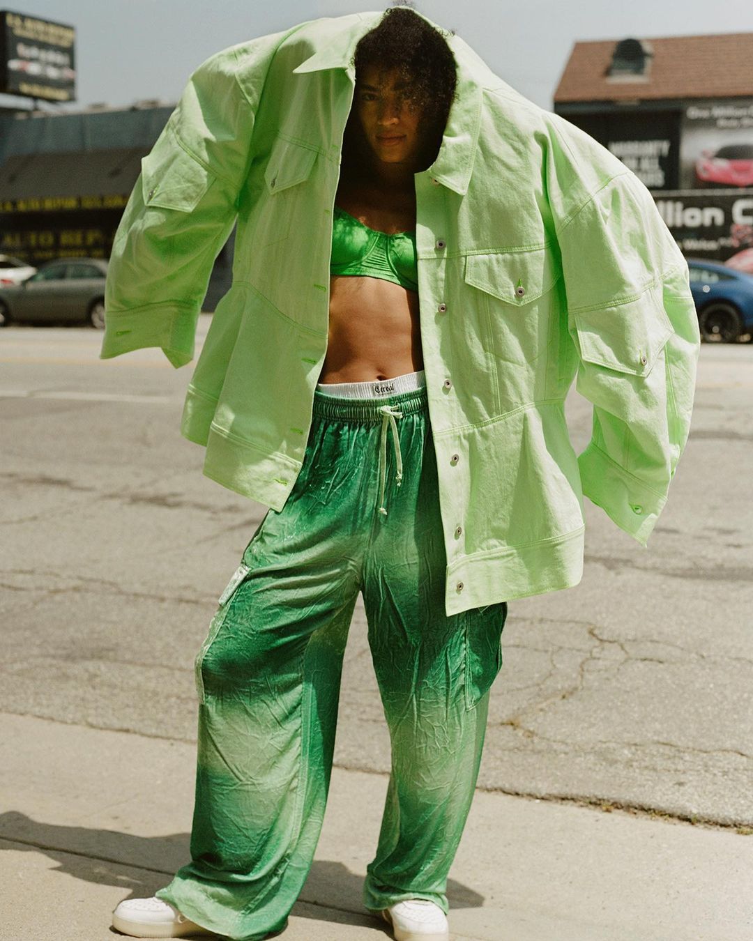 Woman in green bra and oversized jacket and pants stands outdoors on city street with cars and buildings in the background on sunny day.