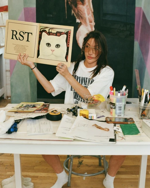 Woman in a white t-shirt sitting at a cluttered table holding a book with a cat illustration in a room with artwork.