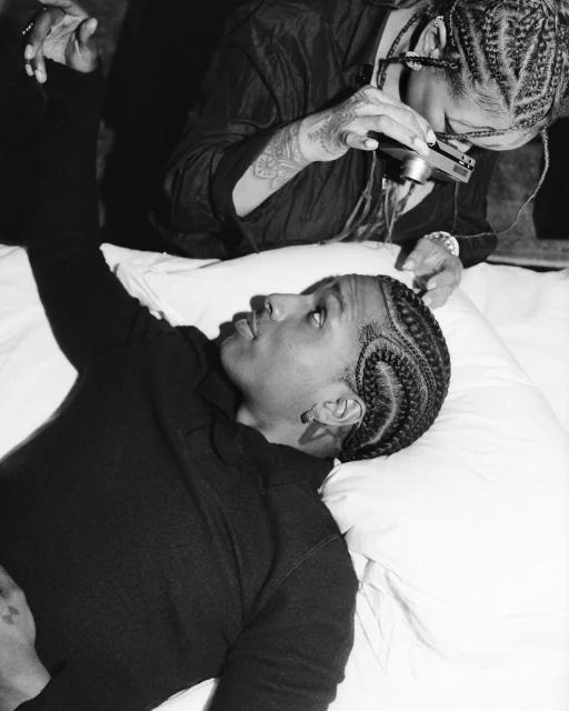 A black-and-white shot captures a young man reclining on a white surface as a person with braids takes a picture with a digital camera.