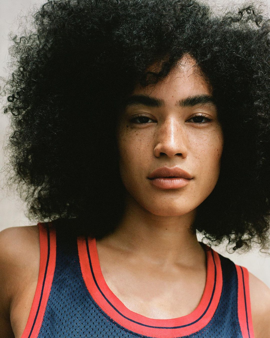 Close-up portrait of a young adult female with freckles and dark, curly hair wearing a sporty mesh tank top in an indoor setting.