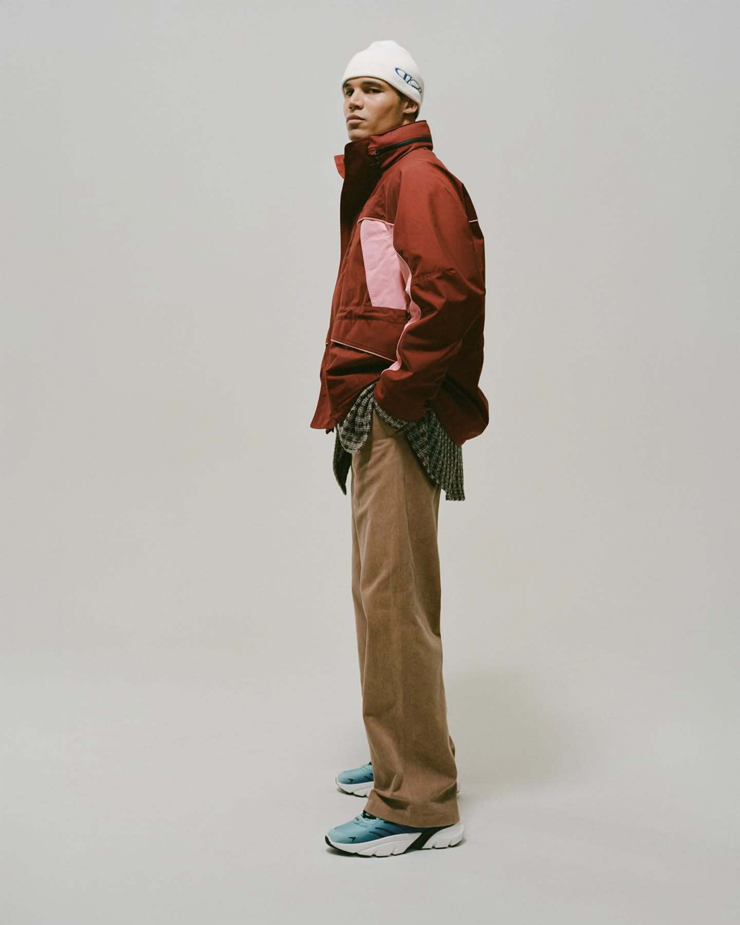 A young adult male model stands in a photo studio wearing a maroon jacket, light brown pants, blue sneakers, and a white beanie.