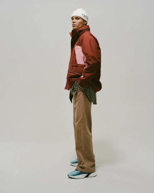 A young adult male model stands in a photo studio wearing a maroon jacket, light brown pants, blue sneakers, and a white beanie.