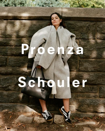 Woman in an oversized light grey coat holding a black handbag and wearing sneakers against a stone wall background.