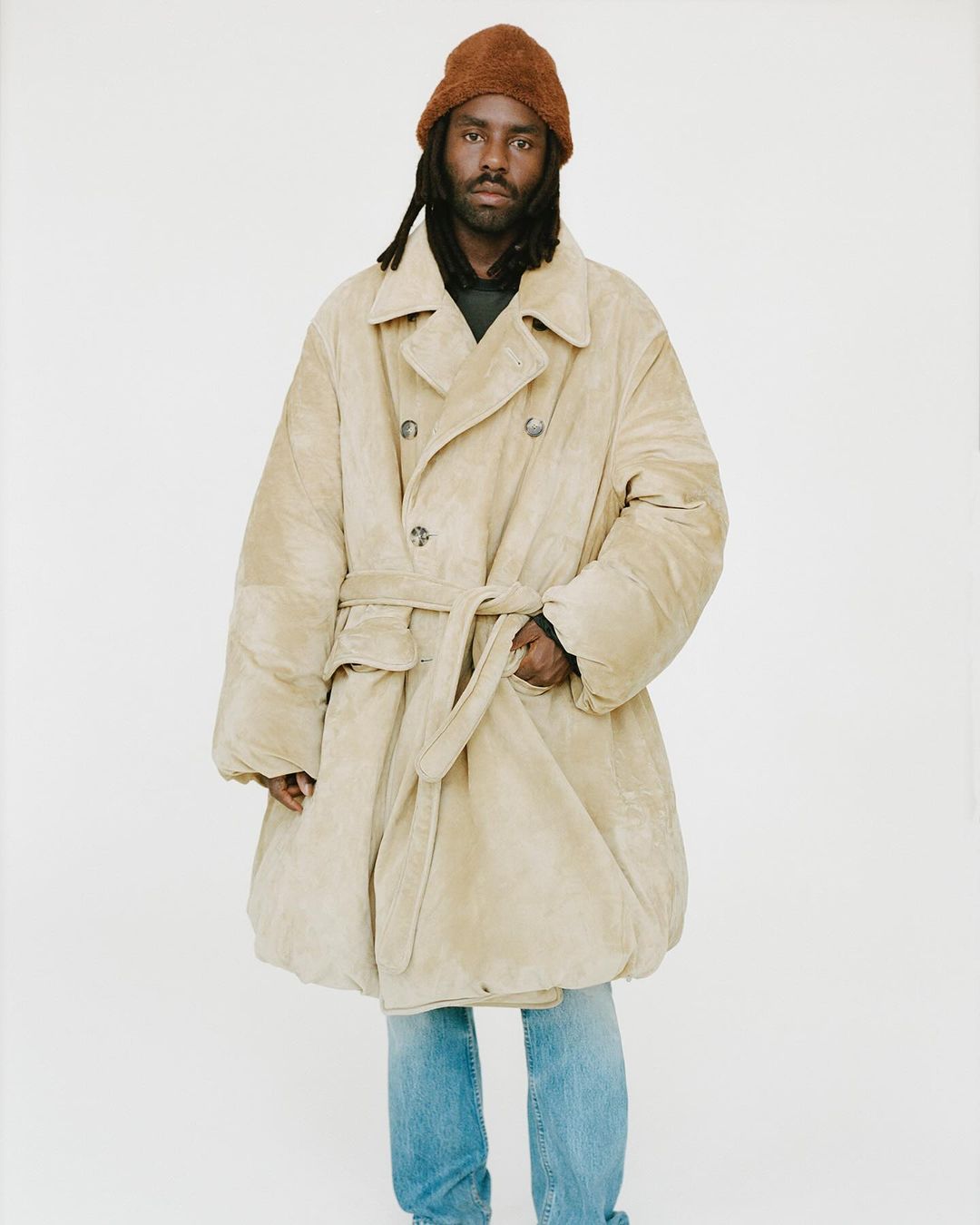 Adult male poses in a stylish oversized coat with a belt and beanie in front of a neutral backdrop, showcasing modern fashion.