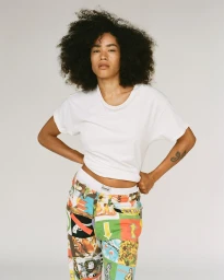 Portrait of a woman with dark curly hair wearing a white t-shirt and colourful patterned trousers against a plain white background