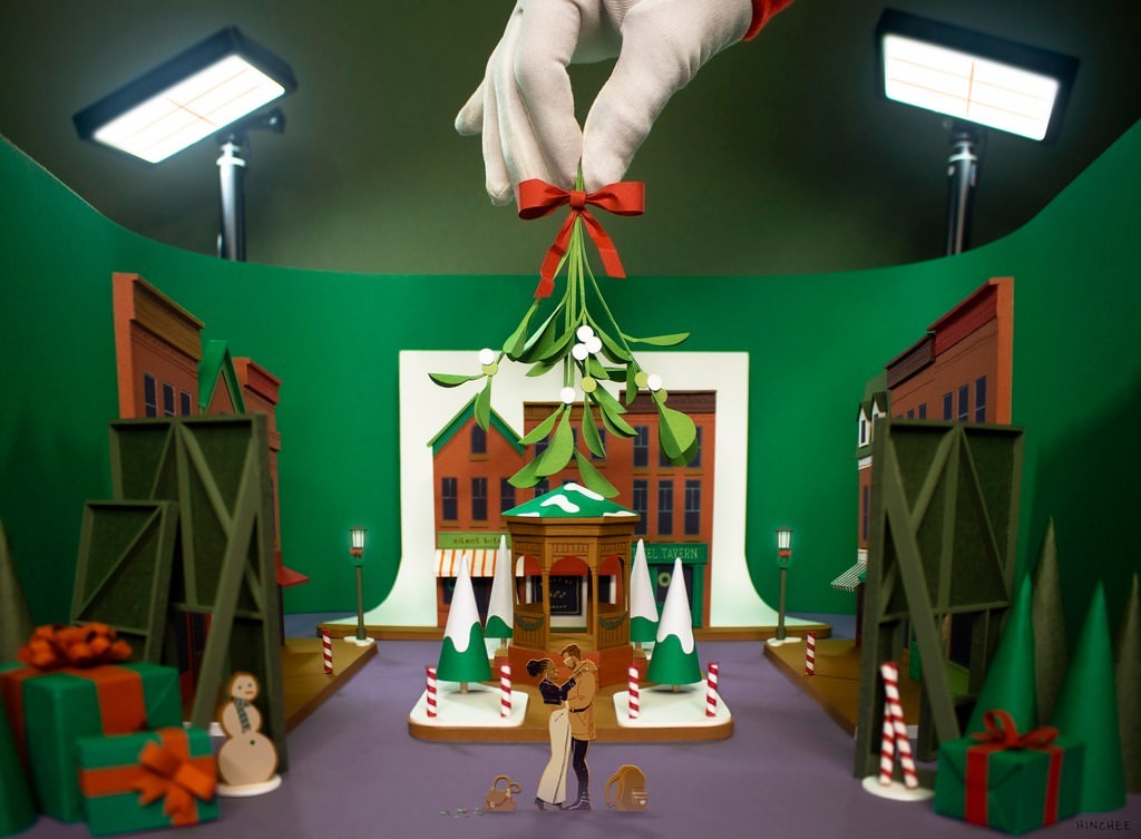 Stop-motion animation shows a miniature couple kissing under mistletoe held by a hand in a green and white glove on a wintery city street.