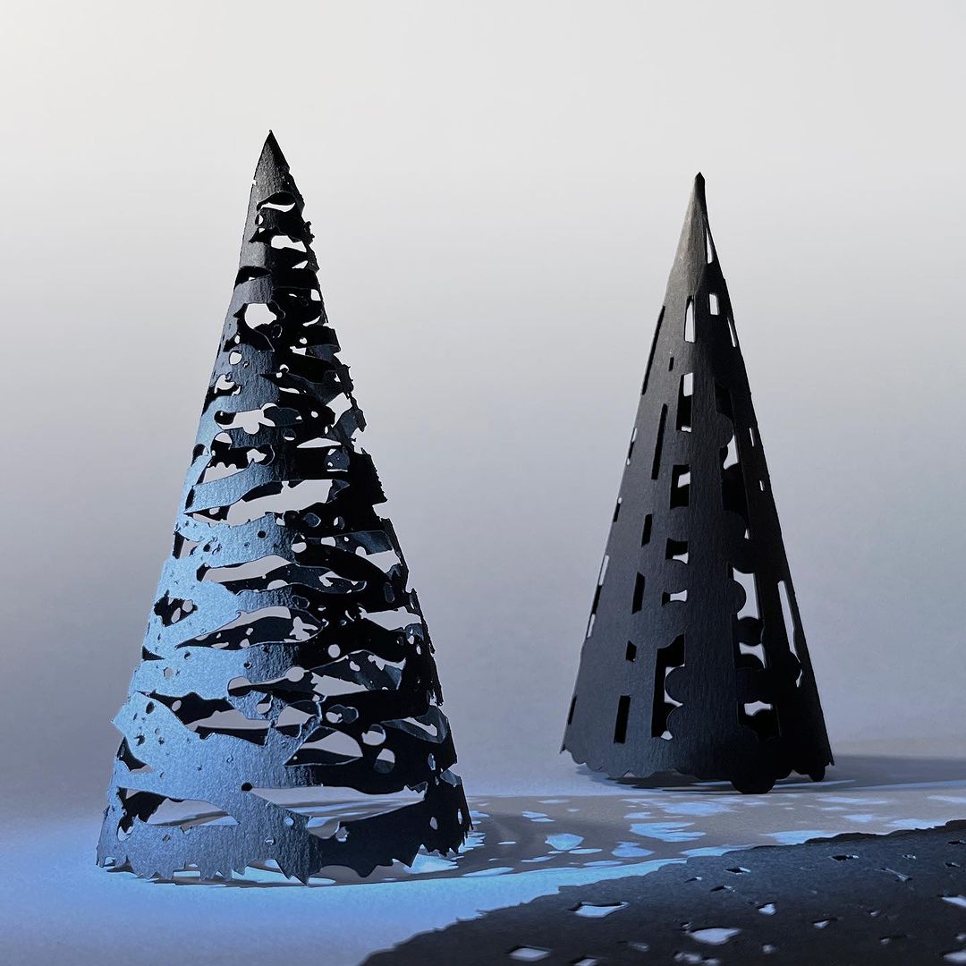 Two dark cut-paper trees stand on a light surface. Abstract cutout patterns create shadows, giving a graphic and artistic paper-art design.