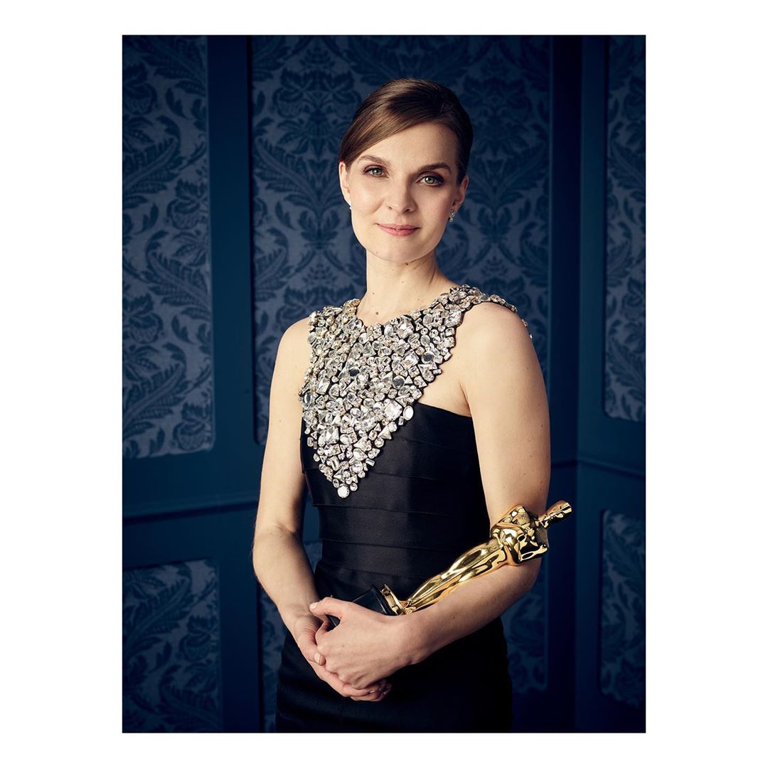 Woman in a black dress with jeweled collar, holding a golden statuette, posing against a dark blue patterned backdrop in a medium shot.