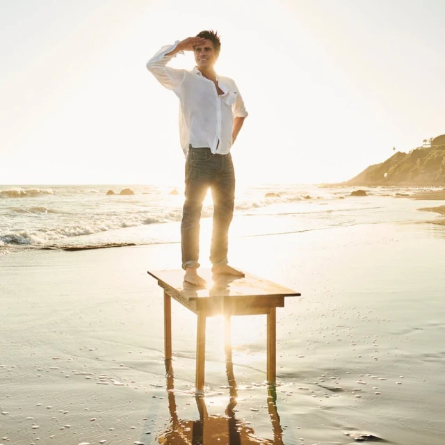 A man standing on a wooden table at a beach, shielding his eyes from the sun. The background includes ocean waves and a bright sky.